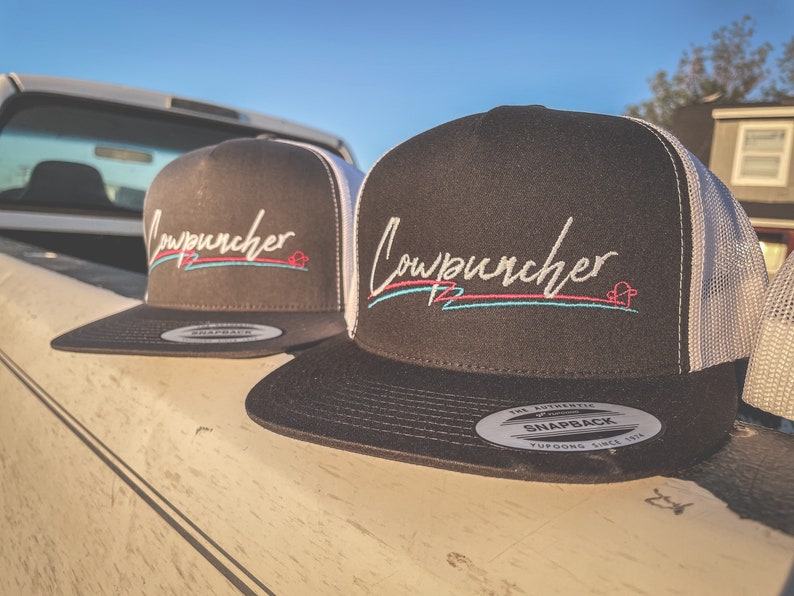 Pu&ograve; includere: Due cappellini da camionista neri con retro in rete bianca. I cappellini presentano la parola "Cowpuncher" ricamata in un font corsivo, con un design rosa e blu sottostante. I cappellini sono appoggiati su una superficie colorata chiara.