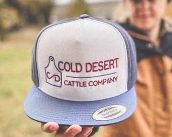 Może przedstawiać: Biało-niebieska czapka trucker z haftowanym logo w kolorze czerwonym i niebieskim, na którym widnieje napis "COLD DESERT CATTLE COMPANY". Logo zawiera stylizowaną literę "D" w kółku.