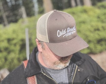 May include: A brown and beige mesh trucker hat with a white embroidered logo that says "Cold Desert Clothing Co."