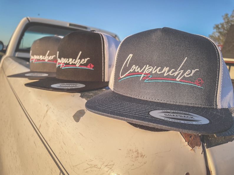 Pu&ograve; includere: Tre cappelli snapback con la scritta "Cowpuncher" ricamata in bianco, con accenti rossi e turchese. I cappelli sono neri, antracite e neri con retro in rete bianca. I cappelli sono appoggiati su una superficie usurata.