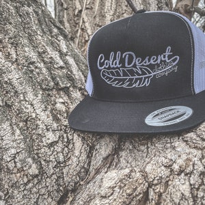 May include: A black and white trucker hat with a white mesh back. The hat has an embroidered logo that says "Cold Desert Clothing Company" with a feather design.
