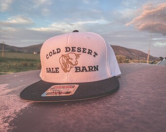 Może przedstawiać: Biało-czarna czapka typu snapback z płaskim daszkiem z napisem "COLD DESERT SALE BARN" i grafiką byka. Czapka ma czarny daszek i metkę na daszku. Tło przedstawia zachód słońca nad wiejskim krajobrazem.