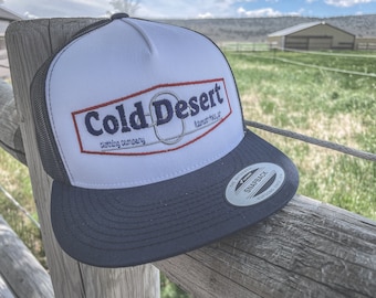 May include: A white and navy blue trucker hat with a red, white, and blue embroidered logo that says "Cold Desert Clothing Company Klamath Falls, OR". The hat has a black mesh back and a snap closure.