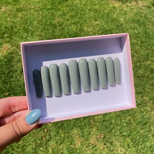 May include: A set of ten green and gray press-on nails in a pink box. The nails are arranged in a row and are all the same size and shape.
