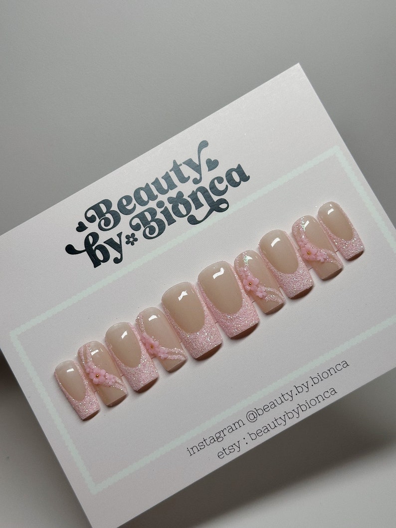 May include: Pink and white glitter press-on nails with a delicate floral design. The nails are decorated with small pink flowers and a thin line of glitter. The nails are arranged in a row on a white background. Instagram: @beauty.by.bionca Etsy: beautybybionca