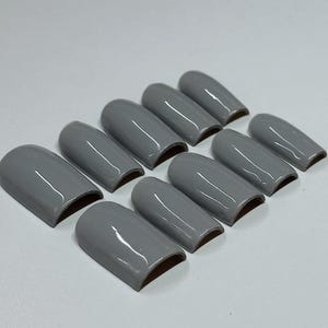 May include: A set of ten gray, glossy, artificial nails arranged on a white surface. Each nail has a rounded shape and a smooth, reflective surface. The nails are a solid, neutral gray color.