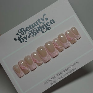 May include: Pink and white press-on nails with glitter and small pink flowers. Instagram: @beauty.by.bionca Etsy: beautybybionca