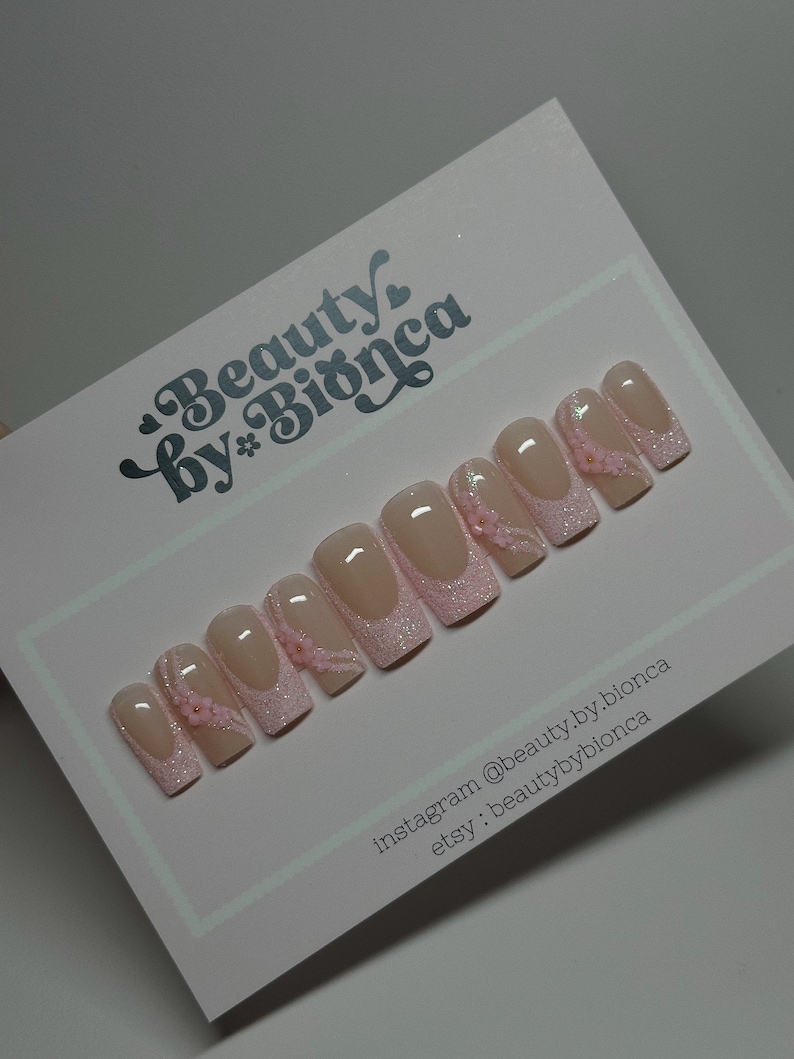 May include: Pink and white press-on nails with glitter and small pink flowers. Instagram: @beauty.by.bionca Etsy: beautybybionca