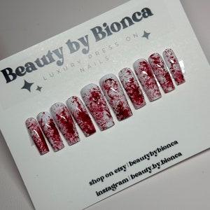 May include: A set of press-on nails with a white base and a splattered red design, resembling blood. The nails are rectangular and arranged on a white card with the text "Beauty by Bionca" and social media information.