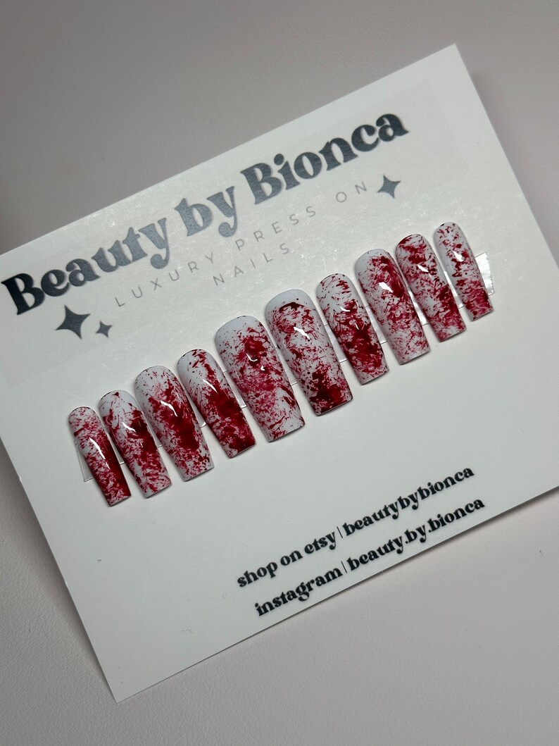 May include: A set of press-on nails with a white base and a splattered red design, resembling blood. The nails are rectangular and arranged on a white card with the text "Beauty by Bionca" and social media information.