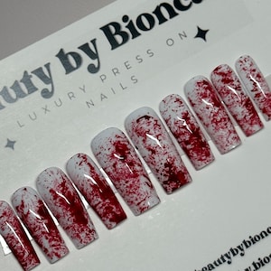 May include: A set of coffin-shaped press-on nails with a white base and a splattered blood-like design. The nails are arranged on a card with the text "beauty by Bionca LUXURY PRESS ON NAILS".