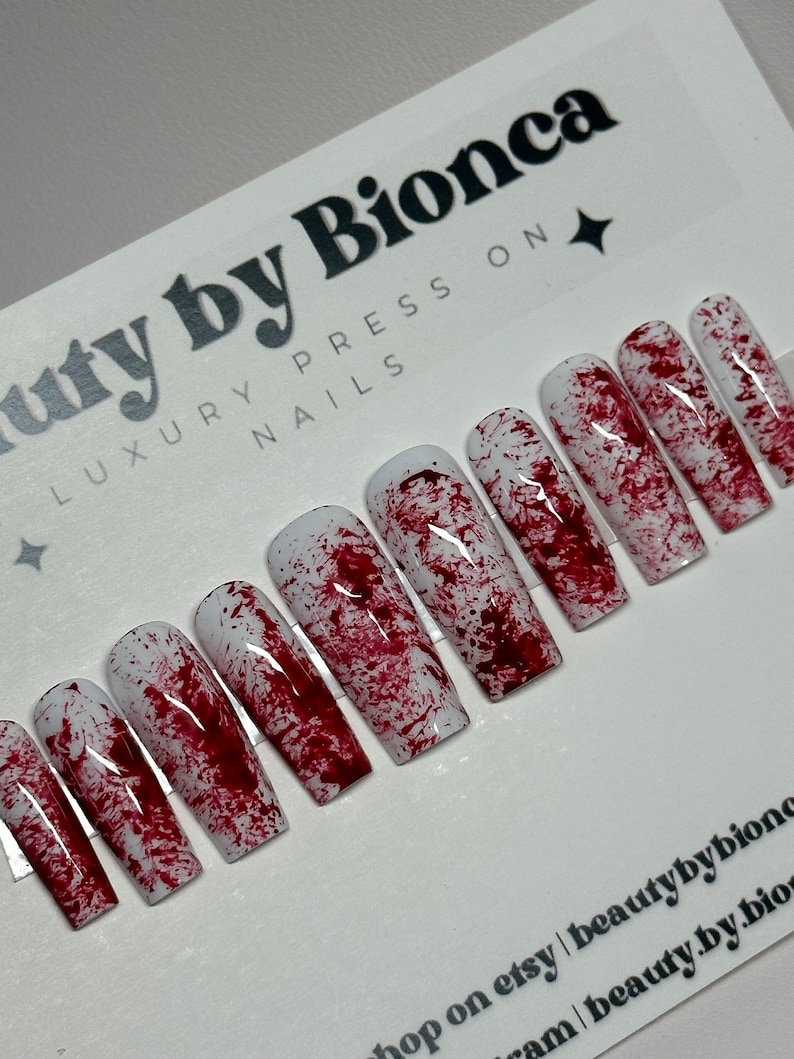 May include: A set of coffin-shaped press-on nails with a white base and a splattered blood-like design. The nails are arranged on a card with the text "beauty by Bionca LUXURY PRESS ON NAILS".