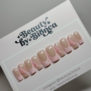May include: Set of pink and beige press-on nails with glitter and small pink flowers. The nails are arranged in a row on a white card with the text "Beauty by* Bianca" and social media handles.
