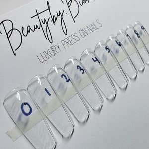 May include: Clear, rectangular press-on nail samples are arranged in a row, each labeled with a number from 0 to 9. The nails are taped to a white card with the text "Beauty by Bianca LUXURY PRESS ON NAILS" in black script.