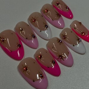 May include: Set of ten almond-shaped artificial nails. Each nail features a nude base with a pink or white tip. Embellishments include small, sparkling red and gold accents, and a few gold dots.