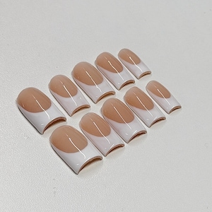 May include: A set of ten artificial nails with a classic French manicure design. Each nail features a natural-toned base with a white tip. The nails are arranged on a white surface, showcasing their shape and style.