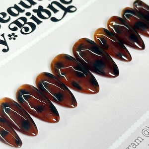 May include: A set of ten brown and black tortoise shell press-on nails. The nails are long and have a pointed shape.