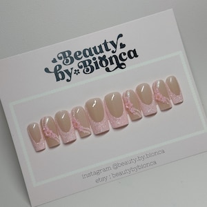 May include: Pink and white press-on nails with glitter and small pink flowers. The nails are displayed on a white card with the text "Beauty by Bianca" and social media information.