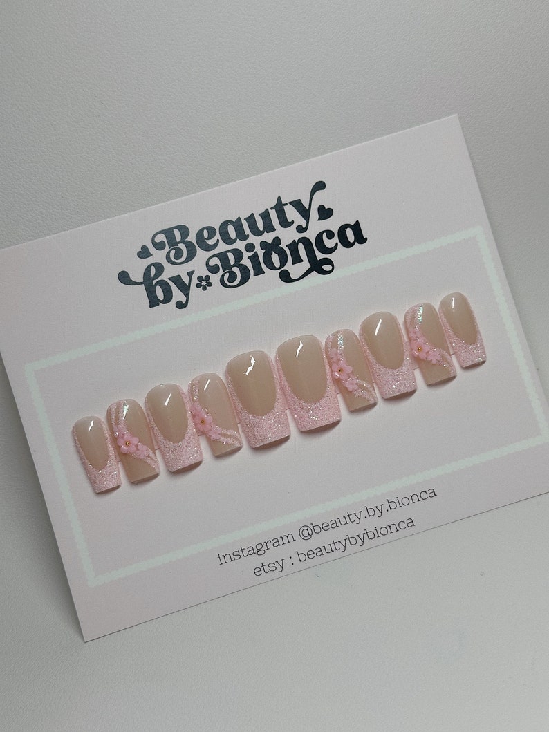 May include: Pink and white press-on nails with glitter and small pink flowers. The nails are displayed on a white card with the text "Beauty by Bianca" and social media information.