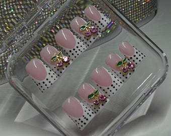 May include: A set of press-on nails in a clear plastic case. The nails are oval-shaped with a light pink base and white tips. Each nail features a gold-colored cherry charm with green leaves and pink gems. The background is white with black polka dots.