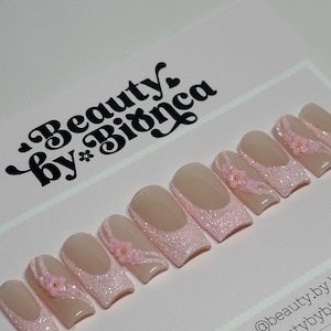 May include: Pink and nude short square-shaped press-on nails with glitter and small pink flowers. The nails are arranged in a row on a pink surface. The text "Beauty by Bianca" is in the top left corner of the image. The text "instagram @beauty.by.bianca etsy: beautybybianca" is in the bottom right corner of the image.