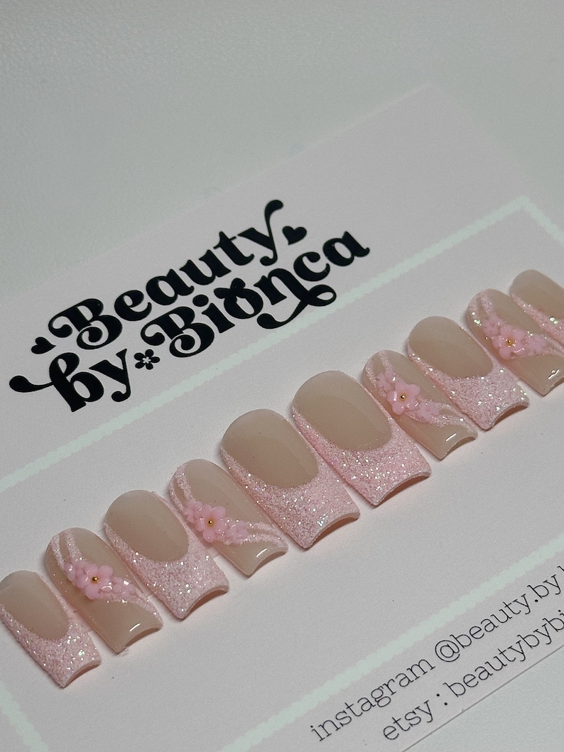 May include: Pink and nude short square-shaped press-on nails with glitter and small pink flowers. The nails are arranged in a row on a pink surface. The text "Beauty by Bianca" is in the top left corner of the image. The text "instagram @beauty.by.bianca etsy: beautybybianca" is in the bottom right corner of the image.