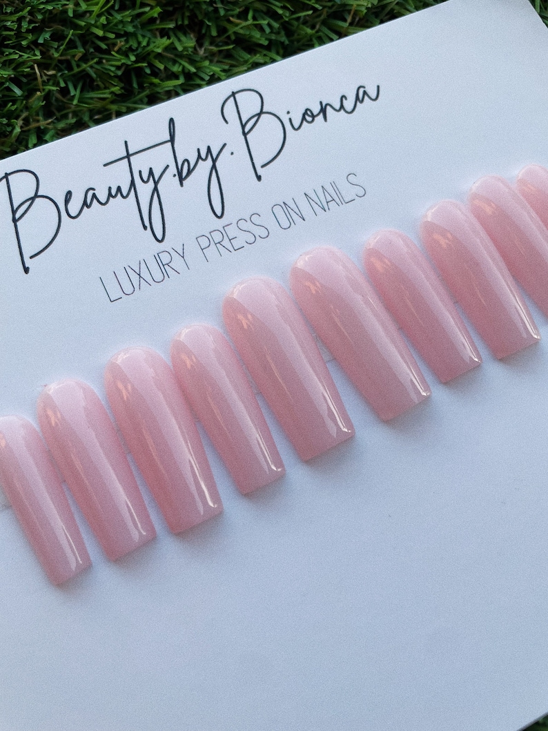 May include: A set of ten long, light pink press-on nails. The nails are arranged in a row on a white surface. The text "Beauty by Bionca LUXURY PRESS ON NAILS" is visible at the top of the image.