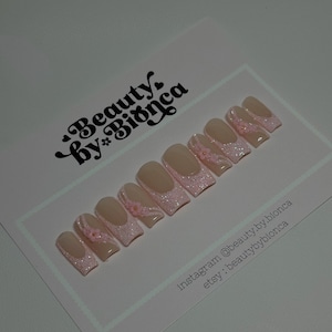 May include: Set of pink and white press-on nails with glitter and small pink flowers. The nails are displayed on a white card with the text "Beauty by Bianca" and social media handles.