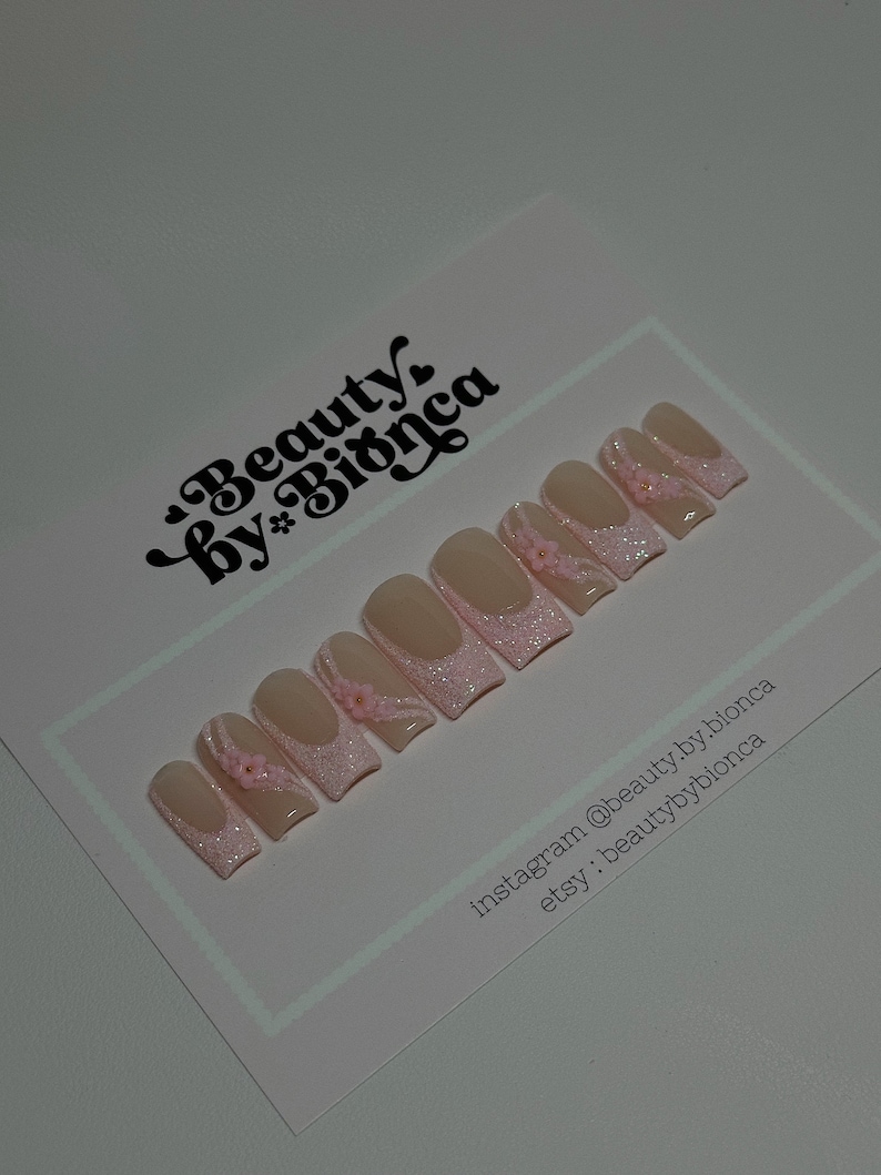 May include: Set of pink and white press-on nails with glitter and small pink flowers. The nails are displayed on a white card with the text "Beauty by Bianca" and social media handles.