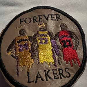May include: A round, brown leather patch with black stitching. The patch features three basketball players in yellow, gold, and red jerseys with the text "FOREVER LAKERS" around the edge.