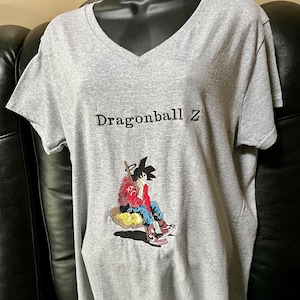 May include: Gray V-neck t-shirt with a graphic print of a cartoon character from Dragonball Z. The character is sitting on a cloud with a staff in his hand.