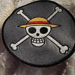 May include: A black and white embroidered patch with a skull wearing a straw hat with a red band. The skull is surrounded by two crossed bones.