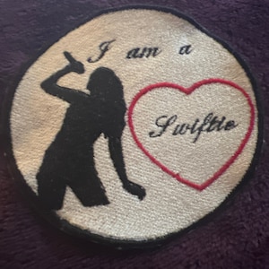 May include: A white, round patch with black embroidery of a woman singing into a microphone. The text "I am a Swiftie" is embroidered in black and red on the patch.