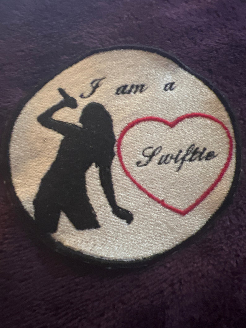 May include: A white, round patch with black embroidery of a woman singing into a microphone. The text "I am a Swiftie" is embroidered in black and red on the patch.