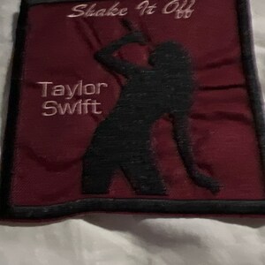 May include: A maroon fabric patch with black embroidered edging. The patch features a black silhouette of a woman singing with the text "Shake It Off" and "Taylor Swift" in white embroidery.