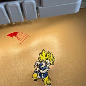 May include: Embroidered patch featuring a cartoon character with spiky yellow hair, blue and white clothing, and yellow pants. The patch is being created by an embroidery machine. A red design is also being embroidered.