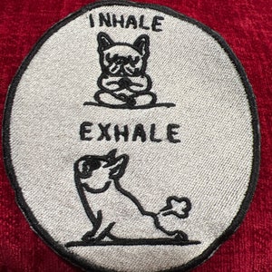 May include: A white oval patch with black embroidery of two dogs. The top dog is sitting with its paws folded in front of it and the text "INHALE" above it. The bottom dog is lying down with its tail curled up and the text "EXHALE" above it.