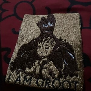 May include: A brown and beige embroidered patch featuring a character from the Guardians of the Galaxy movie franchise. The character is depicted with outstretched arms and the text "I AM GROOT" is embroidered below.