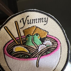 May include: Embroidered patch with a white bowl of ramen noodles, two chopsticks, a yellow egg, and two green frogs. The bowl has a pink rim and the text "Yummy" is written in black cursive above the bowl.