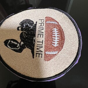 May include: A beige and brown embroidered patch with a black silhouette of a football player holding a football. The text "PRIME TIME" is written vertically on the patch.