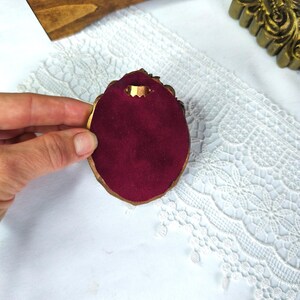 May include: A small, ornate, gold-colored jewelry display with a deep red velvet interior. The display is held against a white lace background, with a wooden box and gold-colored frame in the background.