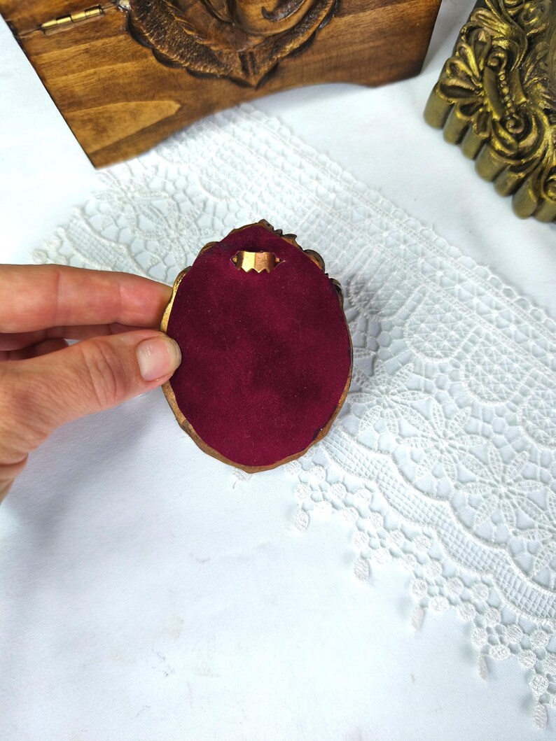 May include: A small, ornate, gold-colored jewelry display with a deep red velvet interior. The display is held against a white lace background, with a wooden box and gold-colored frame in the background.