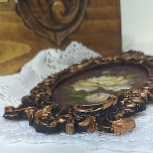 May include: A small, ornate, oval-shaped picture frame with a black and gold finish. The frame is decorated with intricate floral designs and has a white flower painting inside.