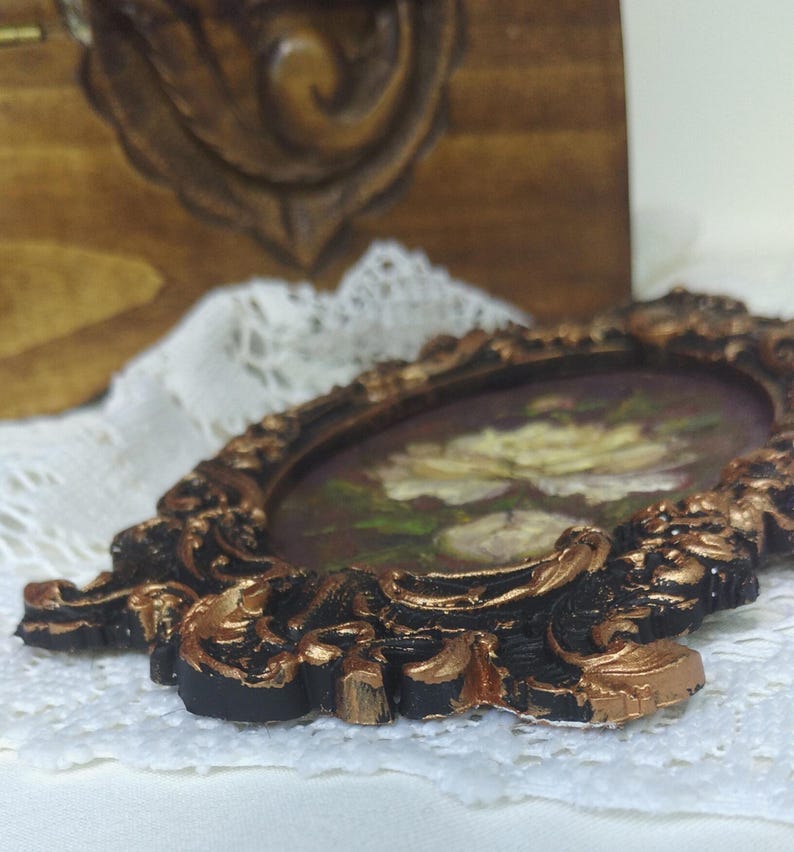 May include: A small, ornate, oval-shaped picture frame with a black and gold finish. The frame is decorated with intricate floral designs and has a white flower painting inside.