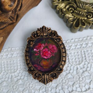 May include: An oval-shaped, bronze-colored frame with an oil painting of pink roses in a vase. The painting is set against a dark background. The frame has ornate detailing and is placed on a white lace surface.