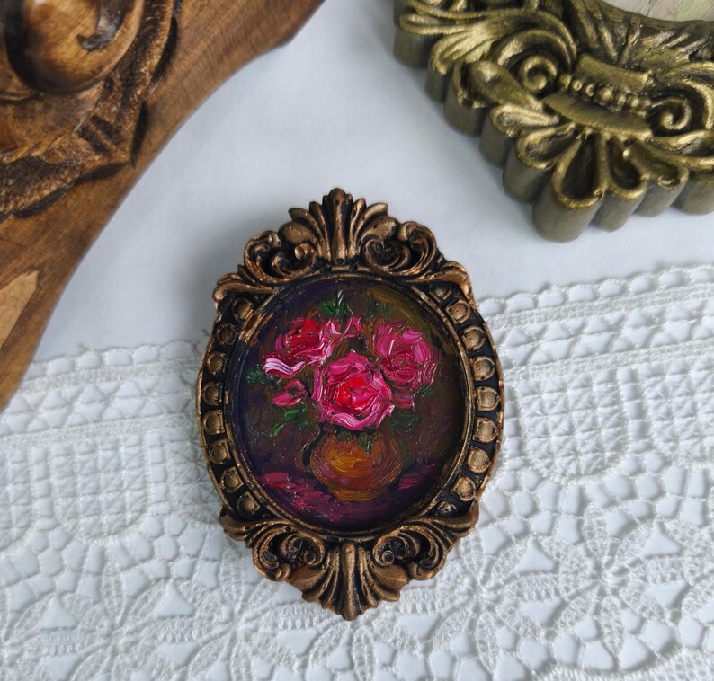 May include: An oval-shaped, bronze-colored frame with an oil painting of pink roses in a vase. The painting is set against a dark background. The frame has ornate detailing and is placed on a white lace surface.