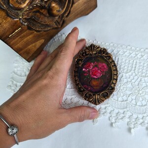May include: An oval-shaped miniature painting in a bronze-colored ornate frame. The painting depicts a vase of pink roses. A carved wooden box is visible in the background. The painting is on a white lace surface.