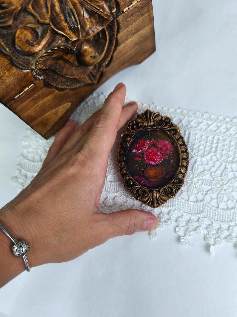 May include: An oval-shaped miniature painting in a bronze-colored ornate frame. The painting depicts a vase of pink roses. A carved wooden box is visible in the background. The painting is on a white lace surface.