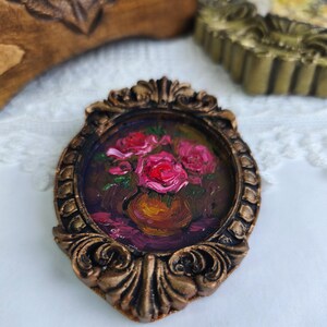 May include: A miniature painting of pink roses in a vase, set within an ornate, bronze-colored oval frame. The artwork features a dark background and is displayed on a white lace surface.