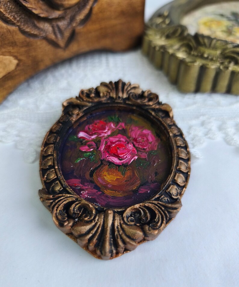 May include: A miniature painting of pink roses in a vase, set within an ornate, bronze-colored oval frame. The artwork features a dark background and is displayed on a white lace surface.